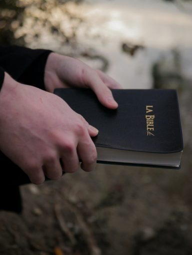 Mains tenant un livre noir intitulé "LA BIBLE" sur fond naturel.