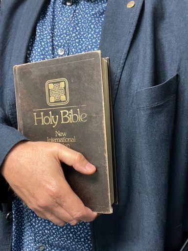 Un homme tient une Bible ouverte près de son torse, vêtu d'une veste bleu foncé.